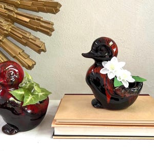 May include: Two ceramic duck planters with a glossy finish. One is filled with greenery, the other with white flowers. The ducks are black with red accents. The word "Canada" is visible on one.