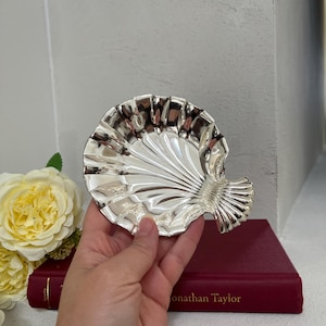 Peut inclure: Un plat en métal argenté brillant en forme de coquille Saint-Jacques. L'objet présente une série de cannelures rayonnantes s'étendant de la base jusqu'au bord festonné, créant une surface réfléchissante avec une finition métallique lisse.