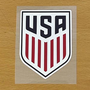 May include: A shield-shaped sticker featuring the letters USA in bold black font at the top, positioned above five vertical red stripes. The entire design is outlined in a thick black border against a white background on a clear adhesive backing.