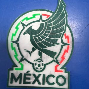May include: A white, green, and red emblem featuring the Mexican coat of arms. The emblem includes a green eagle with a snake in its beak, a soccer ball, and the word "MÉXICO" in green letters. The emblem is set against a blue background.