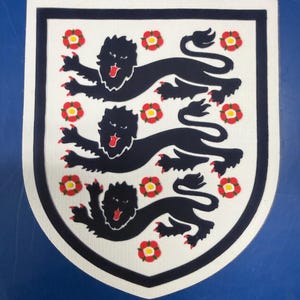 May include: A white shield-shaped emblem with a black border, featuring three black lions passant and six red and yellow Tudor roses. The emblem is likely a crest or patch.