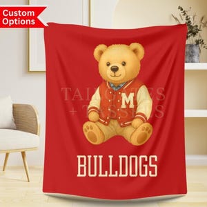 Puede incluir: Manta roja con un oso de peluche que lleva una chaqueta universitaria roja y crema con la letra "M". La palabra "BULLDOGS" está impresa en blanco en la parte inferior. La manta se exhibe en una habitación con una silla y una estantería.