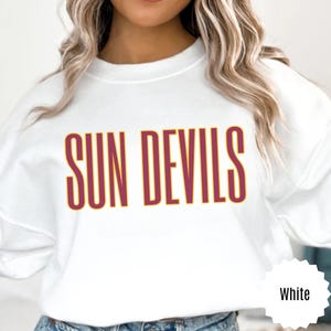 Puede incluir: Sudadera blanca con la inscripción "SUN DEVILS" en letras granates y doradas. La sudadera de cuello redondo tiene mangas largas. El texto está centrado en la parte delantera de la prenda.