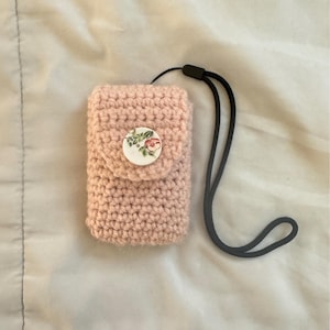 May include: A light pink, crocheted phone case with a floral button closure and a black lanyard. The case is rectangular and appears handmade. The button features a delicate floral design with pink and green accents.
