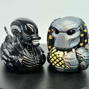 May include: Two detailed figurines of iconic movie monsters. One is a glossy black Alien with a detailed head and body. The other is a Predator, featuring a silver mask, dreadlocks, and armor. Both are on a black surface.