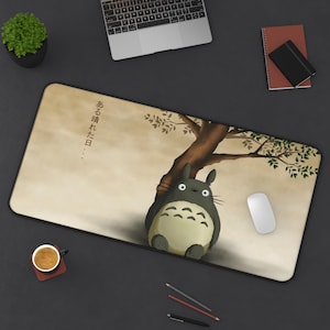 Pode incluir: Tapete de rato retangular com uma ilustração de desenho animado de Totoro do filme de animação Meu Vizinho Totoro. O design inclui uma árvore, o personagem Totoro e texto japonês. O tapete tem uma borda preta e um fundo bege.