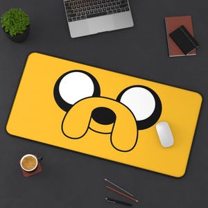 May include: A yellow rectangular mousepad featuring a cartoon dog face with large white eyes and a black nose and mouth. A white computer mouse rests on the right side of the mousepad. A laptop, coffee cup, and pens are also visible.