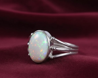 May include: A silver ring featuring an oval opal gemstone. The opal displays a milky white base with iridescent flashes of color. The ring has a split shank design with decorative accents.