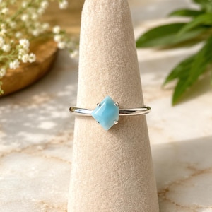 May include: A silver ring featuring a single, kite-shaped, light blue gemstone held in place by four small prongs. The band is smooth and polished, showcasing a clean, metallic finish that complements the soft, opaque blue hue of the stone.