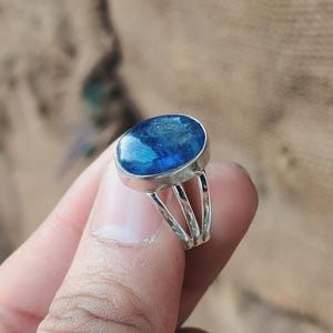 Puede incluir: Un anillo de plata con una gema grande, ovalada y azul oscuro. La gema tiene un aspecto texturizado y el anillo tiene un diseño de caña dividida. El anillo se sostiene sobre un fondo neutro.