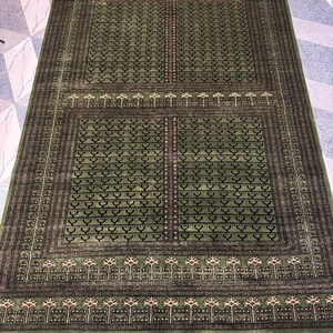 May include: A rectangular green rug with an intricate pattern. The rug features a central field of green with repeating geometric shapes and a border of darker green with a detailed design. The rug has a fringe on the edges.