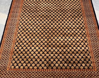 Puede incluir: Una alfombra rectangular con un patrón detallado. La alfombra presenta un borde azul marino oscuro y un campo central de diseños en forma de diamante de color beige y azul marino. La alfombra tiene un borde decorativo con detalles en rojo y beige y una franja.