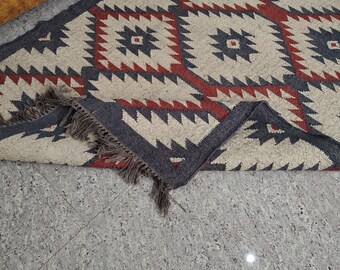 May include: A rectangular rug with a geometric pattern in shades of grey, red, and beige. The rug features a repeating diamond design with fringed edges. The text "Bhadoriya Rugs Home Decor" is visible in the upper left corner.