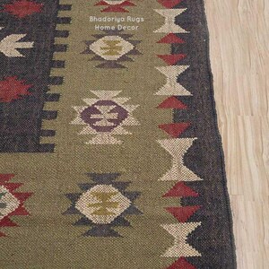 May include: A woven rug featuring a dark charcoal border and a central olive green panel. The rug displays geometric patterns in red, cream, and purple, with the text Bhadoriya Rugs Home Decor printed in white. The bottom edge has a fringe trim.