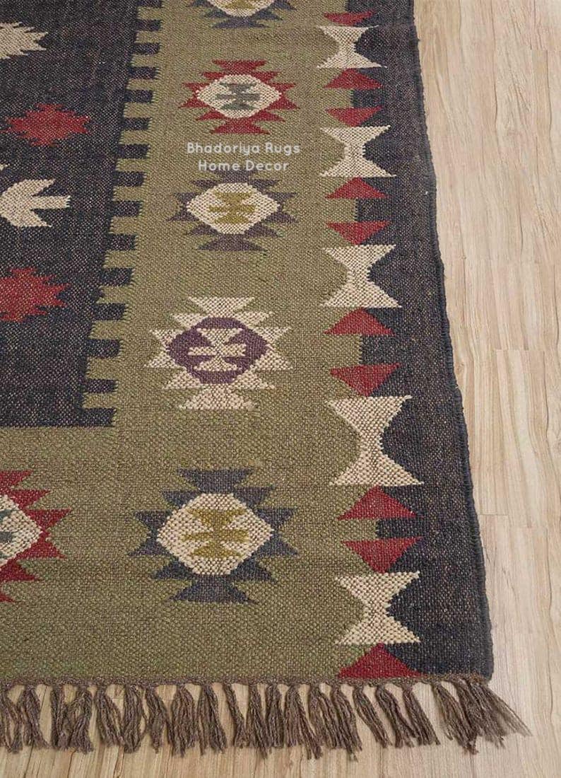 May include: A woven rug featuring a dark charcoal border and a central olive green panel. The rug displays geometric patterns in red, cream, and purple, with the text Bhadoriya Rugs Home Decor printed in white. The bottom edge has a fringe trim.