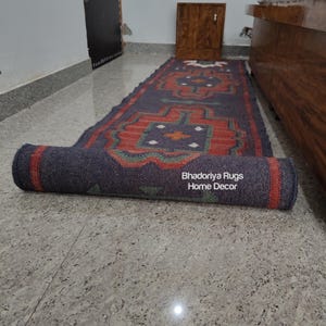 Puede incluir: Una alfombra rectangular enrollada con un patr&oacute;n geom&eacute;trico en rojo, azul y verde. La alfombra est&aacute; parcialmente desenrollada, revelando el dise&ntilde;o. El texto "Bhadoriya Rugs Home Decor" es visible en la parte enrollada. La alfombra est&aacute; sobre un suelo embaldosado.