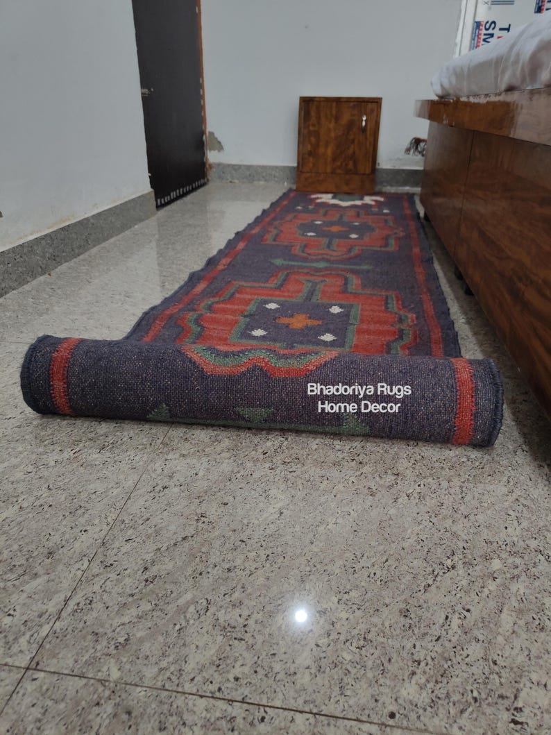 Puede incluir: Una alfombra rectangular enrollada con un patr&oacute;n geom&eacute;trico en rojo, azul y verde. La alfombra est&aacute; parcialmente desenrollada, revelando el dise&ntilde;o. El texto "Bhadoriya Rugs Home Decor" es visible en la parte enrollada. La alfombra est&aacute; sobre un suelo embaldosado.