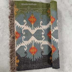 May include: A rolled-up, patterned rug with a green border and fringe. The rug features a geometric design in teal, orange, and white. The text "BhadoriyaRugs" is visible on the top edge.