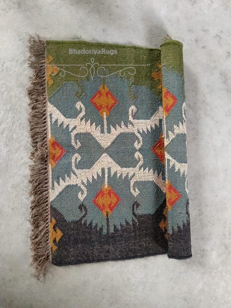 May include: A rolled-up, patterned rug with a green border and fringe. The rug features a geometric design in teal, orange, and white. The text "BhadoriyaRugs" is visible on the top edge.