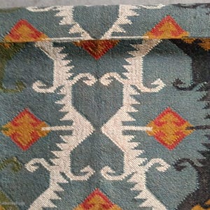 May include: Close-up of a teal rug with a geometric pattern. The design features white, orange, and red diamond shapes and stylized motifs. The rug has a fringed edge.