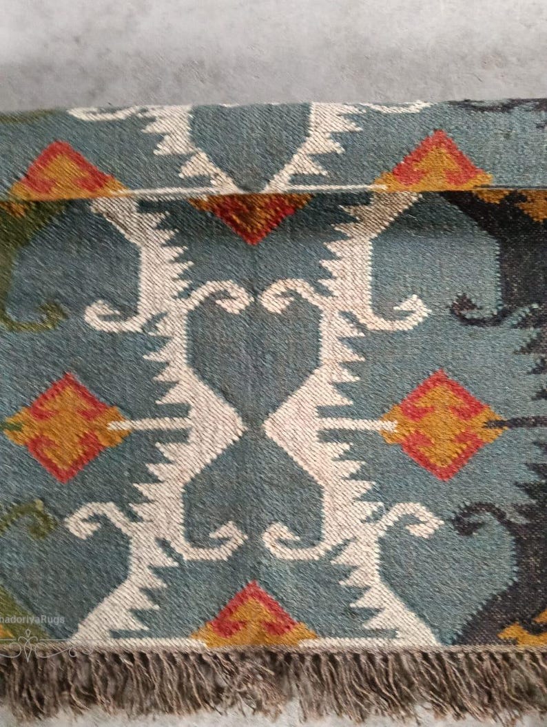 May include: Close-up of a teal rug with a geometric pattern. The design features white, orange, and red diamond shapes and stylized motifs. The rug has a fringed edge.