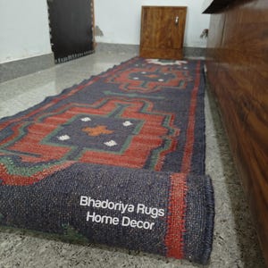 Puede incluir: Una alfombra rectangular enrollada con un patr&oacute;n geom&eacute;trico en tonos de azul, rojo y verde. La alfombra tiene un color base azul oscuro con un dise&ntilde;o repetitivo en rojo y verde. El texto "Bhadoriya Rugs Home Decor" es visible en el borde enrollado.