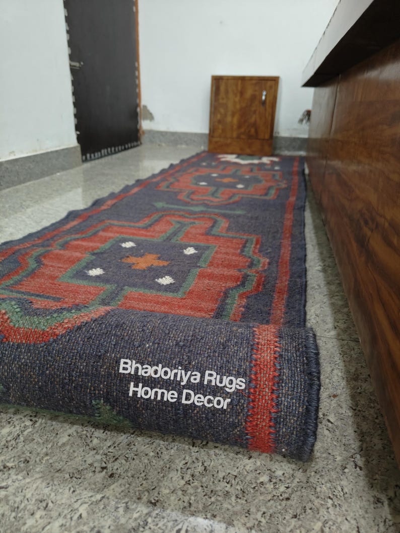 Puede incluir: Una alfombra rectangular enrollada con un patr&oacute;n geom&eacute;trico en tonos de azul, rojo y verde. La alfombra tiene un color base azul oscuro con un dise&ntilde;o repetitivo en rojo y verde. El texto "Bhadoriya Rugs Home Decor" es visible en el borde enrollado.