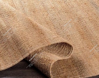 May include: A rolled-up, beige jute rug. The rug has a woven texture and a natural, earthy tone. The edges of the rug are visible, showing the thickness and construction of the material. The rug is on a dark wooden surface.