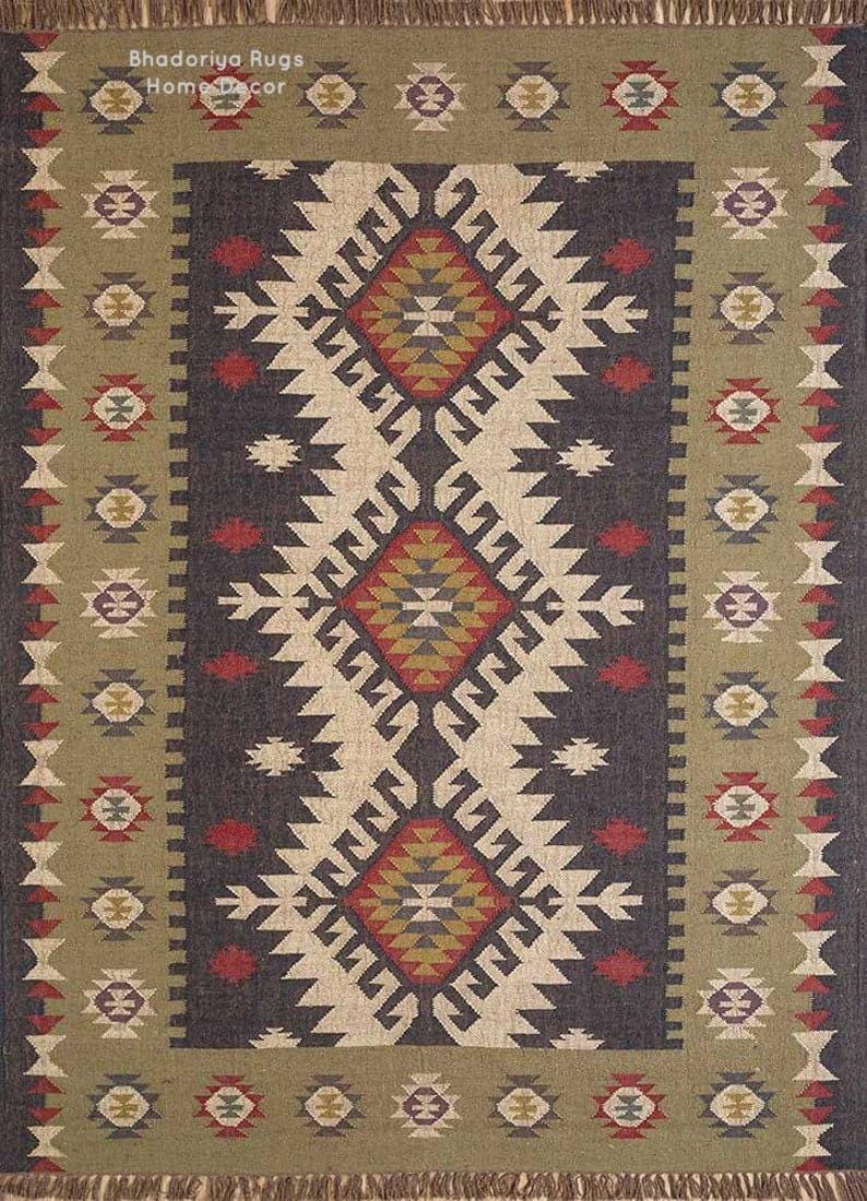 May include: A rectangular area rug featuring a dark charcoal central panel with three large diamond motifs in red, yellow, and white. The border is olive green with repeating geometric shapes and fringe on the top and bottom edges. Text reads Bhadoriya Rugs Home Decor.