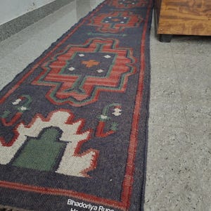 Puede incluir: Una alfombra larga y rectangular con un fondo azul oscuro y patrones geom&eacute;tricos en rojo, verde y blanco. La alfombra tiene un borde con flecos y el texto "Bhadoriya Rugs Home Decor" es visible.
