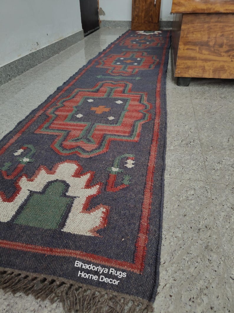 Puede incluir: Una alfombra larga y rectangular con un fondo azul oscuro y patrones geom&eacute;tricos en rojo, verde y blanco. La alfombra tiene un borde con flecos y el texto "Bhadoriya Rugs Home Decor" es visible.