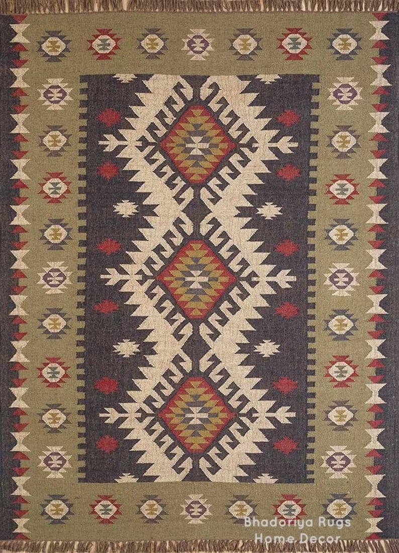 May include: A rectangular area rug featuring a dark charcoal center panel with three large, stacked diamond motifs in red, gold, and cream. The border is olive green with repeating geometric shapes and a fringed edge. Text reads Bhadoriya Rugs Home Decor.