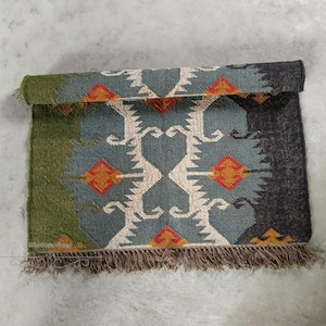 May include: A rolled-up, rectangular rug with a geometric pattern. The rug features a central design in teal, cream, and orange, framed by green and dark gray sections. Brown fringe trims the bottom edge.