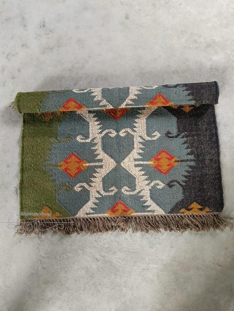 May include: A rolled-up, rectangular rug with a geometric pattern. The rug features a central design in teal, cream, and orange, framed by green and dark gray sections. Brown fringe trims the bottom edge.