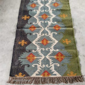 May include: Rectangular rug with a geometric pattern in shades of blue, green, and orange. The rug has a fringed edge and a symmetrical design featuring diamond and arrow shapes. The rug is approximately 36 inches long and 24 inches wide.