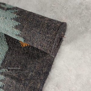 May include: Close-up of a rolled-up, textured rug with a dark brown and teal pattern. The rug's edge is visible, showcasing its thickness and weave. The design includes a mix of brown, teal, and gold colors, creating a rustic aesthetic.