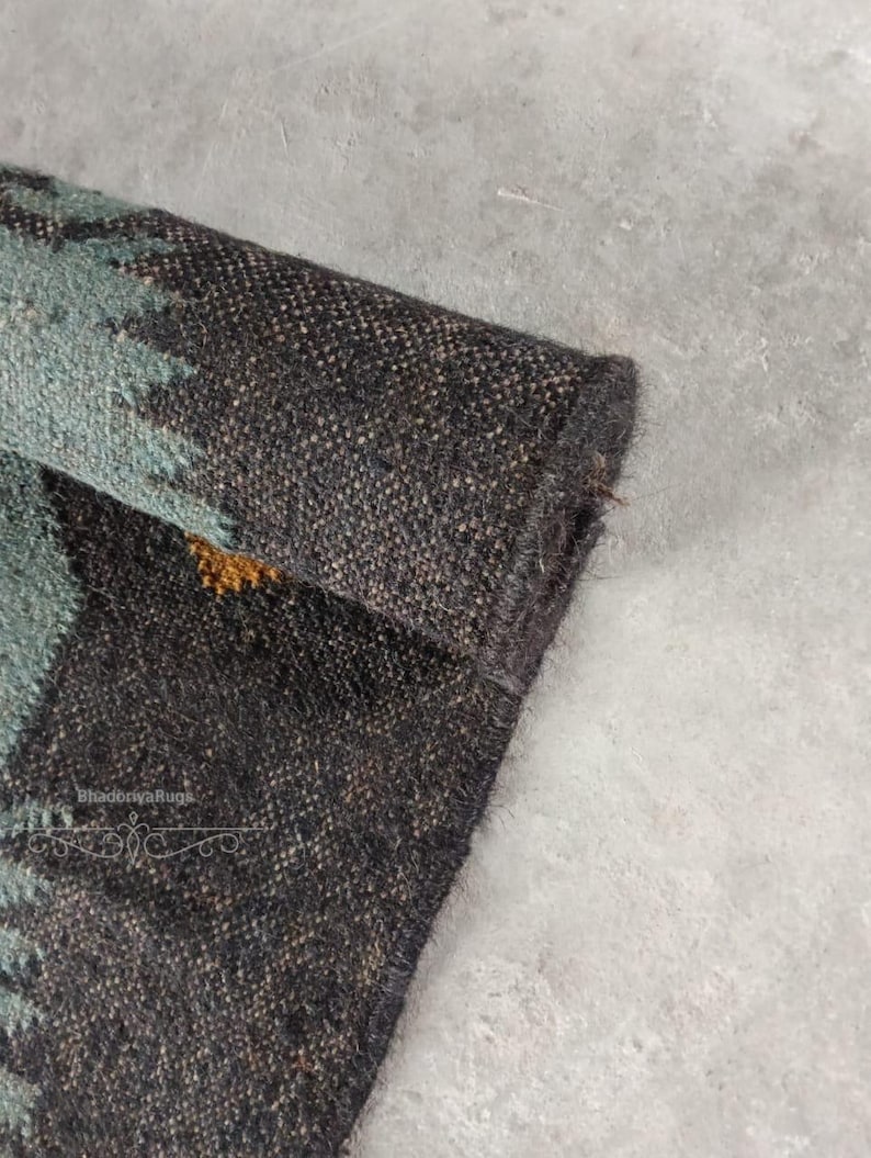 May include: Close-up of a rolled-up, textured rug with a dark brown and teal pattern. The rug's edge is visible, showcasing its thickness and weave. The design includes a mix of brown, teal, and gold colors, creating a rustic aesthetic.