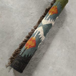 May include: Rolled-up area rug with a teal, orange, and white geometric pattern. The rug has a dark green border and a dark brown fringe. The rug is on a gray concrete surface.