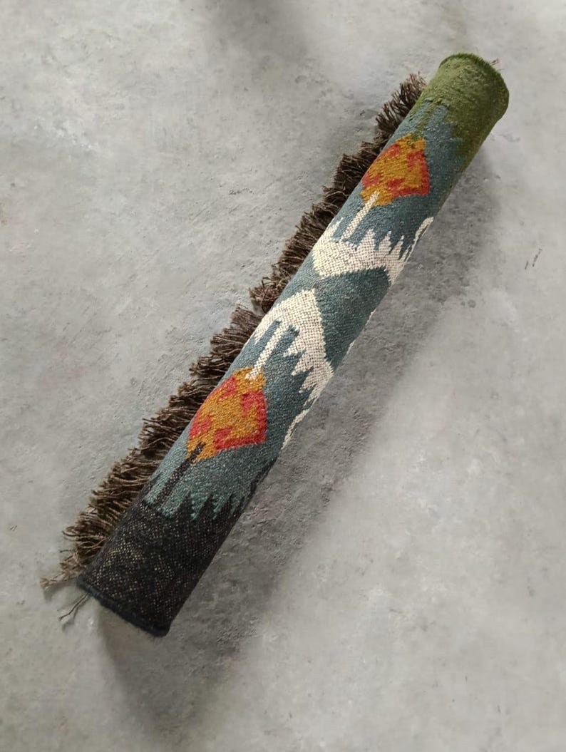 May include: Rolled-up area rug with a teal, orange, and white geometric pattern. The rug has a dark green border and a dark brown fringe. The rug is on a gray concrete surface.