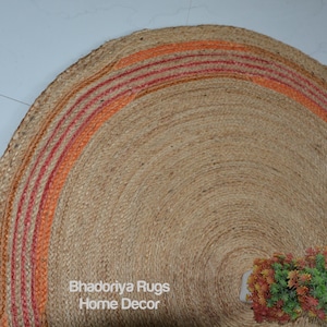 May include: A round jute rug with a natural tan color and an orange and red striped border. The rug has a woven texture and a small decorative plant in the lower right corner. The text "Bhadoriya Rugs Home Decor" is visible.