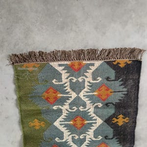 May include: A rectangular rug with a geometric pattern. The rug features a central design in teal, white, and orange, framed by sections of green and dark gray. The edges are finished with a brown fringe.