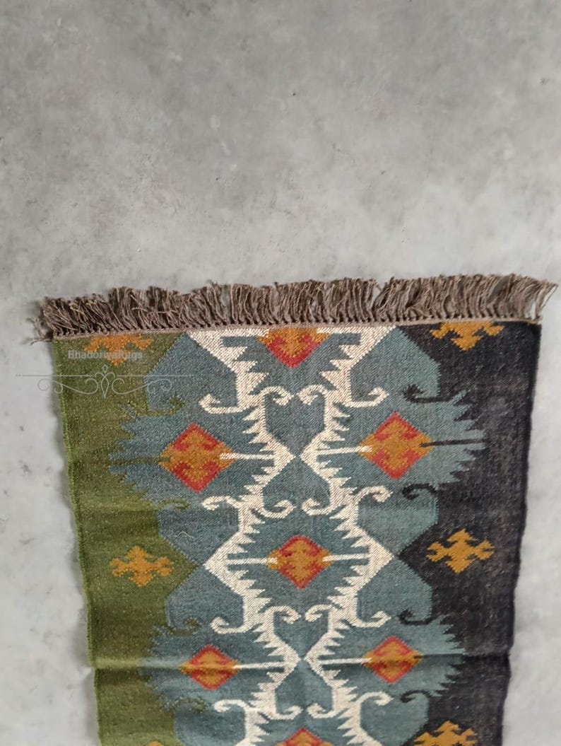 May include: A rectangular rug with a geometric pattern. The rug features a central design in teal, white, and orange, framed by sections of green and dark gray. The edges are finished with a brown fringe.