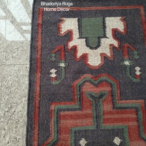 Puede incluir: Una alfombra rectangular con un patr&oacute;n geom&eacute;trico en tonos azul oscuro, rojo, verde y blanco. La alfombra tiene un borde con flecos y el texto "Bhadoriya Rugs Home Decor" es visible.