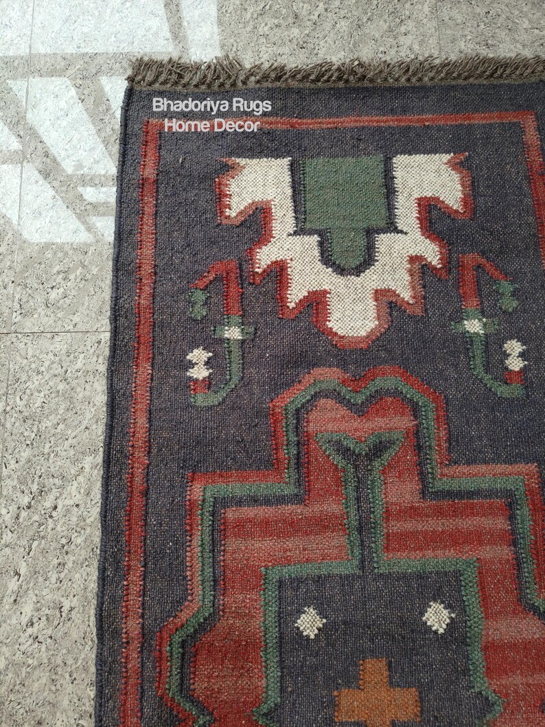 Puede incluir: Una alfombra rectangular con un patr&oacute;n geom&eacute;trico en tonos azul oscuro, rojo, verde y blanco. La alfombra tiene un borde con flecos y el texto "Bhadoriya Rugs Home Decor" es visible.