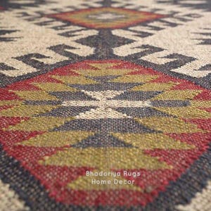 May include: A woven rug featuring a repeating geometric pattern in shades of cream, charcoal black, olive green, and deep red. The textile displays a textured surface with a central diamond motif and the text Bhadoriya Rugs Home Decor printed.