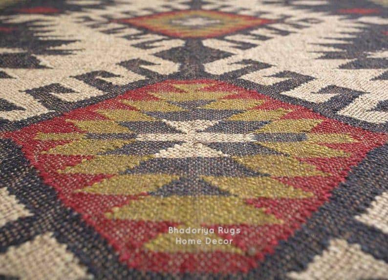 May include: A woven rug featuring a repeating geometric pattern in shades of cream, charcoal black, olive green, and deep red. The textile displays a textured surface with a central diamond motif and the text Bhadoriya Rugs Home Decor printed.