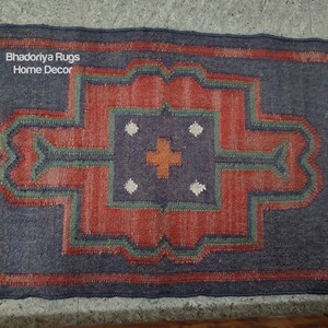 Puede incluir: Una alfombra rectangular con un dise&ntilde;o geom&eacute;trico. La alfombra presenta un patr&oacute;n central rojo y naranja rodeado por un borde azul. El dise&ntilde;o incluye detalles verdes y blancos. El texto "Bhadoriya Rugs Home Decor" es visible.