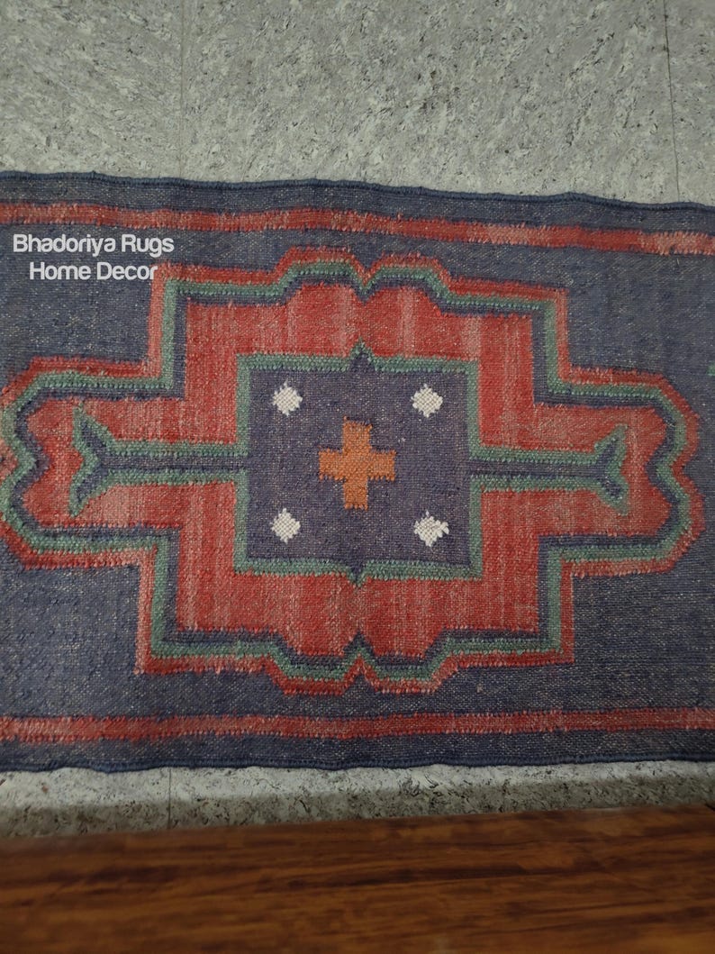 Puede incluir: Una alfombra rectangular con un dise&ntilde;o geom&eacute;trico. La alfombra presenta un patr&oacute;n central rojo y naranja rodeado por un borde azul. El dise&ntilde;o incluye detalles verdes y blancos. El texto "Bhadoriya Rugs Home Decor" es visible.