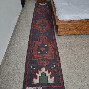 Puede incluir: Una alfombra larga y rectangular con un fondo gris oscuro y un patr&oacute;n geom&eacute;trico repetitivo en rojo, verde y blanco. La alfombra tiene un borde con flecos y el texto "Bhadoriya Rugs Home Decor" es visible.
