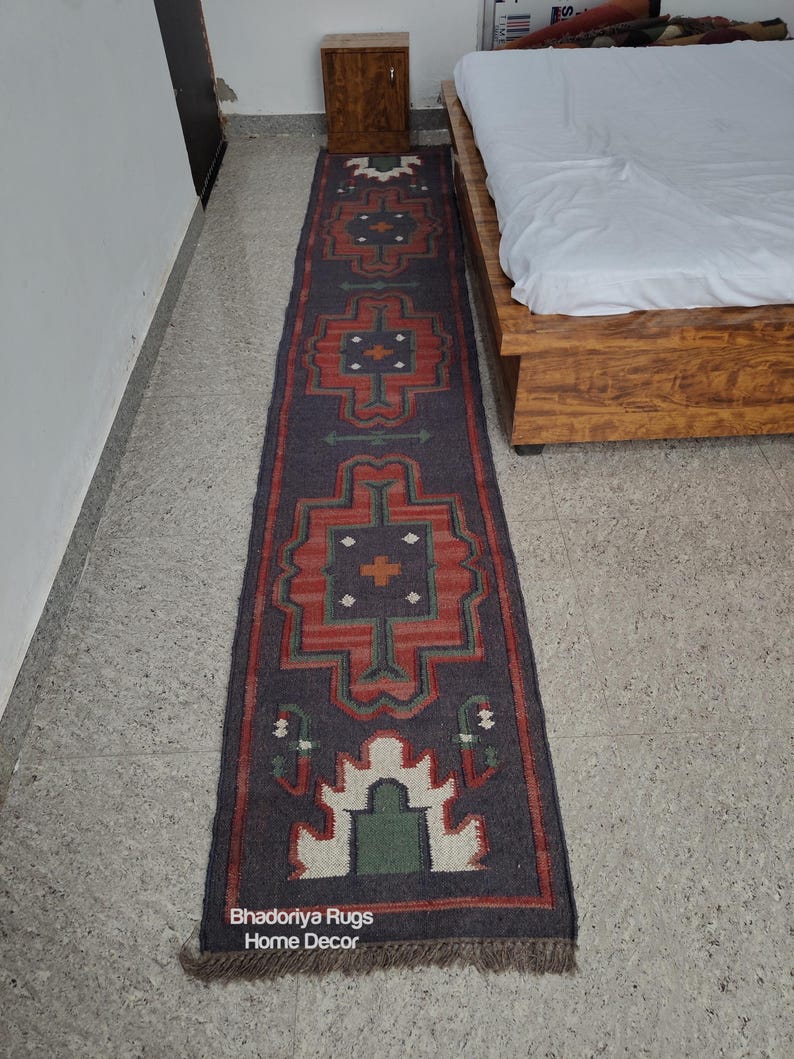 Puede incluir: Una alfombra larga y rectangular con un fondo gris oscuro y un patr&oacute;n geom&eacute;trico repetitivo en rojo, verde y blanco. La alfombra tiene un borde con flecos y el texto "Bhadoriya Rugs Home Decor" es visible.